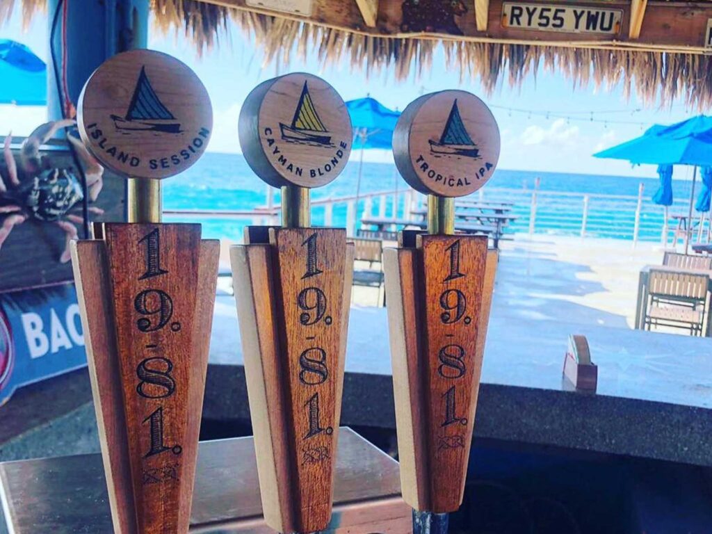 Three beer taps with wooden handles, each featuring a different 19-81 beer logo, against a blurred background of a sunny outdoor patio with blue umbrellas and the ocean