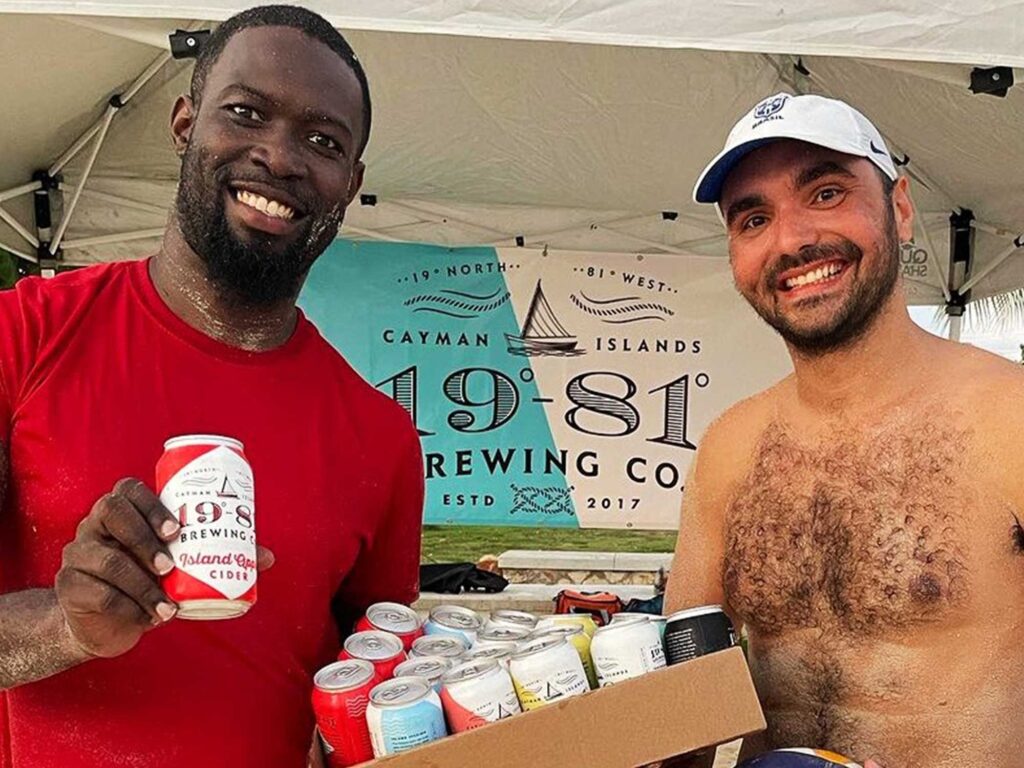 Two men carrying 19-81 beers on a beach in Grand Cayman