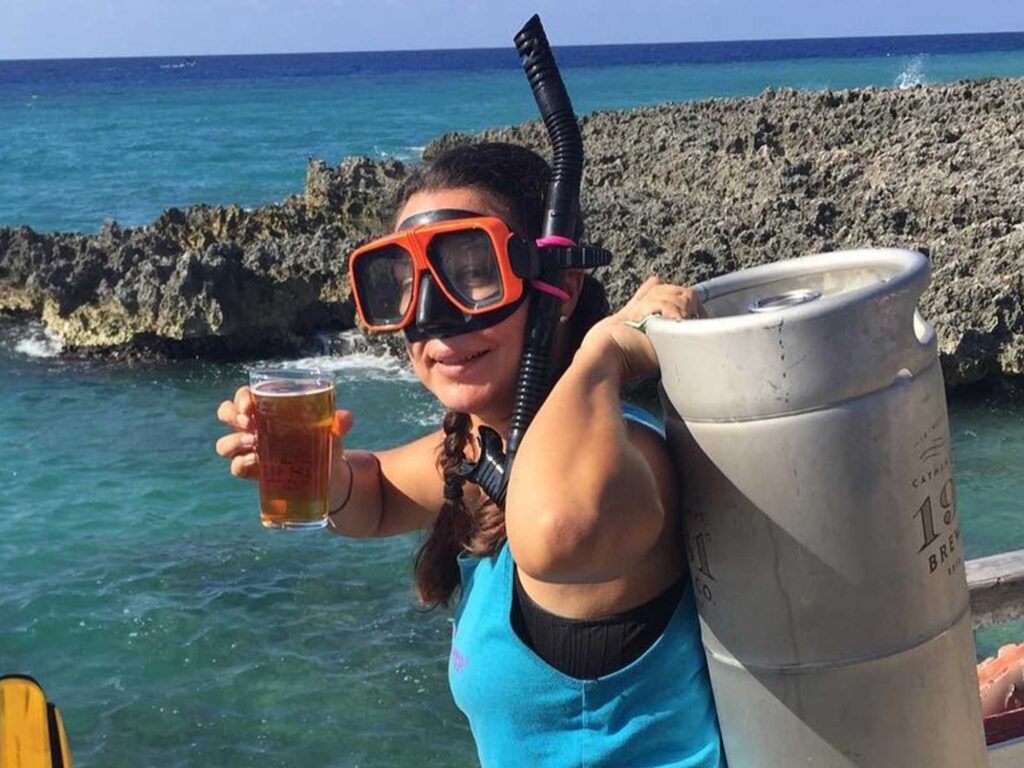 A woman on a beach in the Cayman Islands carrying a 19-81 beer keg, wearing a snorkel mask, and holding a beer